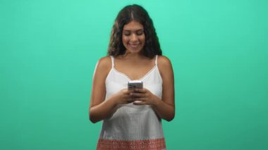 Akıllı telefondan mesaj atan ve stüdyoda elinde yeşil fon ve elbise ile gülümseyen genç İspanyol kadın. Keyif bağlantısı:.