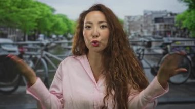 Kadın, kanalın yanında elleri açık omuz silkip jestler yapıyor ve Amsterdam caddesine parketmiş bisikletleri sorguluyor..