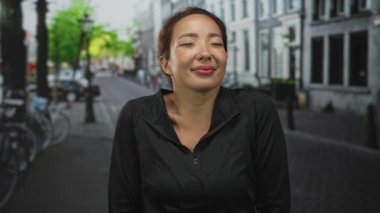 Boynu açıkta bir kadın başını çeviriyor ve sırıtarak gülümsüyor sokakta gözleri kapalı bisiklet ve binalarla dolu; sakin bir memnuniyet.