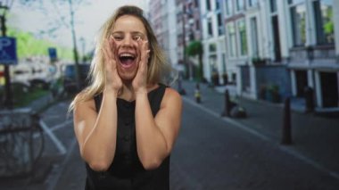 Dar bir şehir caddesinde, kanal binaları ve cesurların yanında bağırarak el ele tutuşan genç sarışın kadın, geniş bir gülümsemeyle; heyecan kutlaması.