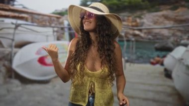 Sarı örgü bluzlu ve güneş şapkalı bir kadın, bir teknenin yanındaki başparmağı işaret ediyor, yüzünü kıvırcık saçlı çerçeveli;.