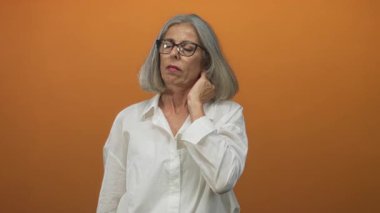 Gri saçlı ve gözlüklü bir kadın boyna dokunuyor ve turuncu stüdyoda elini kaldırıyor..