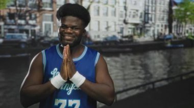 Bir adam, bileklikler ve mavi basketbol forması ile bir caddede, Amsterdam Kanalı 'nın yanında el sıkışıyor; sakin iyimserlik.