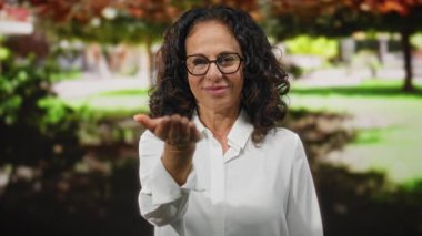 Kıvırcık saçlı ve gözlüklü bir kadın canlı bir açık hava parkı ortamında kameraya doğru elini uzatıyor. Sıcaklık ve dostluk saçıyor..