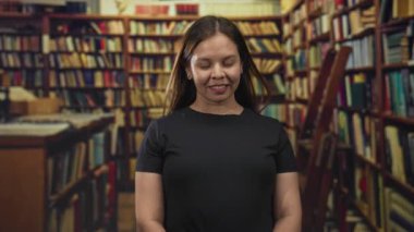 Kütüphane binasındaki kitaplarla çevrili ahşap raflarda dururken gözleri kapalı gülümseyen kadın; sakin yansıma.