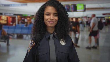 Young hispanic policewoman in uniform with badge extends hand toward camera inside busy airport terminal; friendliness.