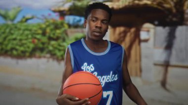 Mavi Los Angeles formalı adam basketbol topunu tutuyor ve güneş ışığıyla aydınlatılmış ev girişinde başını kaşıyor..