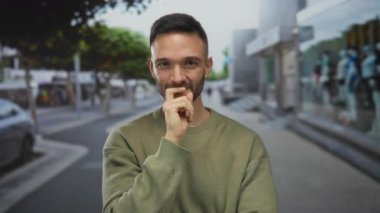 Gündelik süveter giyen genç adam, arka planda ağaçlar ve binalar olan şehir merkezinde gülümsüyor, mutluluk ve kendine güveni somutlaştırıyor..