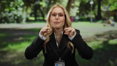 Parktaki kadın, güneşli bir günde arka planda yemyeşil ağaçların arasından süzülen güneş ışığıyla doğal ortamda gözlük ve kimlik kartı takarak anlamlı jestler yapıyor..