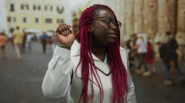 Kırmızı örgülü Afro-Amerikan kadın yoğun bir şehir caddesinde dikkatlice dinliyor, bulanık kentsel arka planla çevrili, açık hava aktivitesi ve etkileşimini tasvir ediyor..