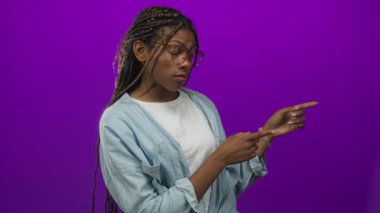 Parmakla işaret eden ve yan bakan bir kadın canlı mor stüdyo ortamında gözlük ve kot gömlek giyer..