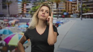 Sarışın genç bir kadın, deniz kıyısındaki bir binanın yanında eli kalçasında bir vneck t-shirt 'le dikilirken parmak tapınağa işaret ediyor..