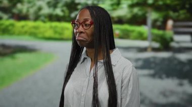 Uzun örgülü saçlı ve gözlüklü bir kadın güneşli bir parkta şaşkın bir ifade sergiliyor..