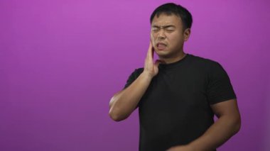 Siyah gömlekli genç adam mor stüdyo ışıklandırmasında diş ağrısı için iki eliyle yanak kavrar..