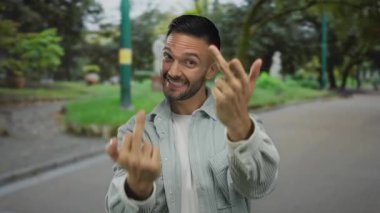 Genç İspanyol bir adam kaba bir jest yapıyor ve yeşil bir parkta gülümsüyor, doğanın ortasında asi ya da kaygısız bir tavır sergiliyor..