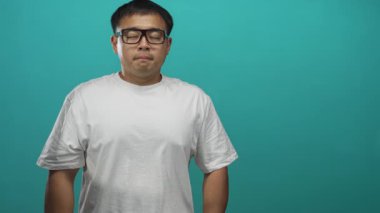 Siyah gözlüklü beyaz tişörtlü adam gözleri yumuşak bir deniz mavisi stüdyo arkaplanına hafifçe kapatır; dinginlik.