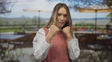 Bir kadın açık teras restoranında neşeli bir şekilde poz veriyor. Şehir manzarası bulanık. Gençlik enerjisi ve gündelik tarzını sergiliyor..