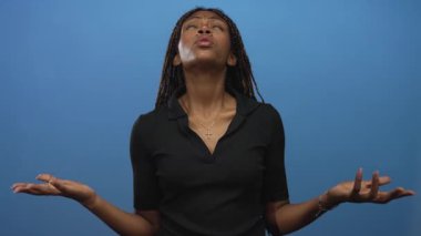 Mavi bir stüdyoda siyah polo ve altın haçlı kolye takan açık avuçlu ve gözleri olan Afro-Amerikalı bir kadın..