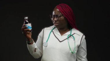 Kırmızı örgülü ve steteskoplu Afro-Amerikan kadın bir şurup şişesi tutuyor ve boğazına dokunuyor, siyah bir arka planda izole edilmiş, boğaz ağrısı için ilaç öneriyor..
