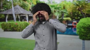 Kıvırcık saçlı bir adam, havuz kenarında çardakta iki elini alnına bastırıyor. Çıplak eller baş ağrısı için gözlerini kapatıyor. Stres yorgunluğu endişesi..