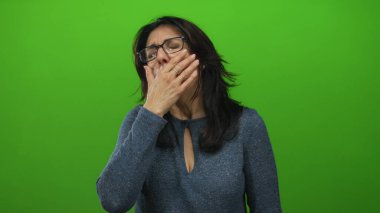 Latin kökenli orta yaşlı bir kadın, yeşil bir stüdyoda ağzı kapalı, gözlüklü, yorgun bir ifade takınıyor..