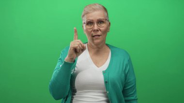 Stüdyoda işaret parmağı yukarı doğru, yeşil ekran karşısında, beyaz v yaka üzerinde gözlük ve deniz mavisi hırka giyen bir kadın..