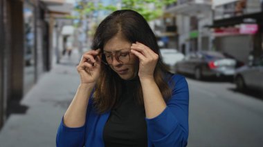 Kadın, mavi ceket ve gözlük takan, stres yorgunluğu çeken şehir merkezindeki caddelerde tapınak basıyor..