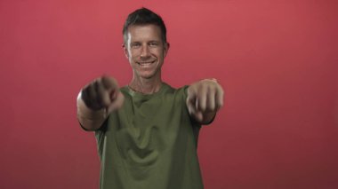 Stüdyoda yeşil tişörtlü bir adam kırmızı arka planda kamerayı işaret ediyor..