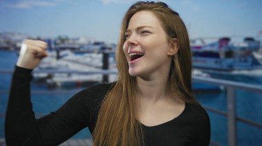 Siyah kazaklı genç kadın liman iskelesinde gözlerini kapatırken yumruğunu sıkıyor; azim zaferi.