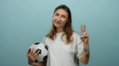 Elinde futbol topu tutan kadın mavi duvara karşı barış işareti gösteriyor. Neşeli duygular ve özgüven ifade ediyor..