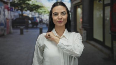 Latin kökenli genç bir kadın, arka planı bulanık, beyaz gömlek giyerken kendine güveni ve gururu yüksek ve gün ışığında gülümseyen şehir sokağındaki kameraya doğru elini uzatıyor..