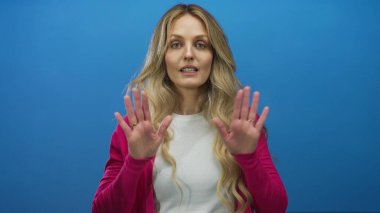 Elleri mavi bir arkaplanda, pembe bir kazak ve beyaz bir gömlek giymiş sakin bir şekilde duran ve izole bir sahneye odaklanan bir kadın..