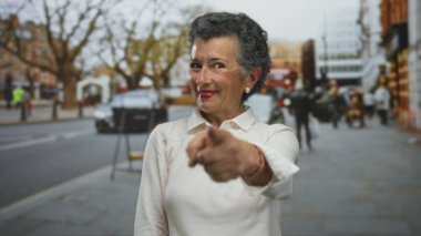 Gri saçlı yaşlı kadın gülümsüyor ve şehir arka planında bulanık bir şehir sokağındaki kameraya bakıyor, hareketli bir kasabada canlı bir açık hava manzarası yakalıyor..