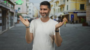 Avuç içleri açık, güneşli bir caddede çıplak elleri ve ön kollarını gösteren bir adam dükkan ve tentelerle dolu, kameraya gülümseyen, sıradan bir eğlence..