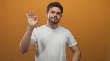Latin asıllı genç adam el işaretini kameraya dönük yapıyor ve turuncu stüdyo ortamında beyaz tişört giyerken rahat bir gülümseme takınıyor..