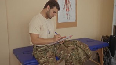 Kamuflaj üniformalı bir adam klinik odasındaki panoya anatomi çizelgesi ve tekerlekli sandalye ile yazıyor..