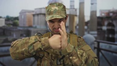 Kamuflaj üniformalı genç İspanyol adam, bina harabelerinde elini kaldırarak bir yumruğu engelliyor; kararlılık.