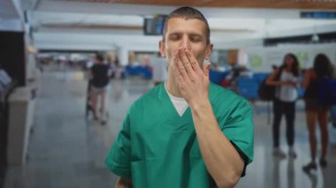 Havalimanı terminalinde gülümseyen ve üfleyen yeşil üniformalı doktor, arka planda bulanık gezginler ve modern mimarisi olan klinik kıyafetli bir erkek var..