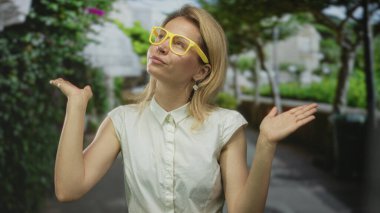 Sarı gözlüklü kadın, yapraklı yeşil ağaçların altında iki elini kaldırıyor. Belirsizlik..
