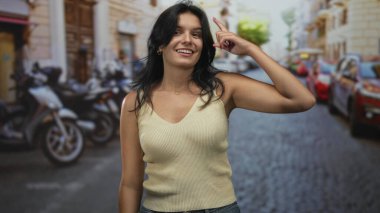 Woman pointing finger to head on a sunlit cobblestone street lined with parked scooters and cars; thinking.