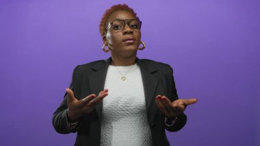 Stüdyoda açık ellerle nefes alan bir kadın gözlük takıyor ve beyaz bluz yerine siyah bir ceket giyiyor..