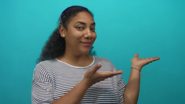 Afrikalı Amerikalı kadın gülümsüyor ve stüdyoda turkuaz duvara karşı çizgili gömlek ile avuç içi açık sunuyor..