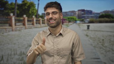 Gözlüklü, bej gömlekli genç gün ışığında sokakta baş parmak işareti yapıyor. Onaylama iyimserliği.