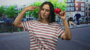 Kanal binalarının yanındaki bir sokakta parmaklarının arasında kırmızı bir kalp tutan bir kadın, çizgili bir tişört giyiyor; umut hayırseverlik farkındalığı.