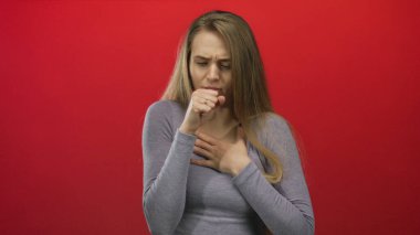 Elini göğsüne koyup kırmızı arka plana doğru öksüren bir kadın, izole edilmiş bir ortamda, günlük gri giysiler içinde sağlık sorunları ve endişelerini dile getiriyor..