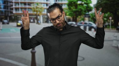 Man making air quotes with both hands and wearing glasses on a busy city street with buildings and parked cars; playful irony.