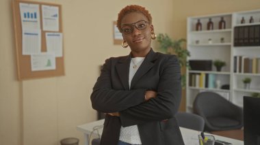 Gözlüklü genç Afrikalı Amerikalı kadın siyah ceketi ilikliyor ve ofis binasında kollarını kavuşturuyor..