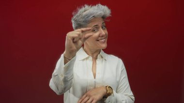 Kadın, beyaz gömlek ve tahta kol saati takarken stüdyoda kırmızı arka plana karşı küçük boy göstermek için parmaklarını çimdikliyor; şüphecilik.