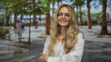 Young blonde woman in a white shirt confidently poses on an outdoor street lined with trees and blurred pedestrians, exuding a sense of urban style and confidence.