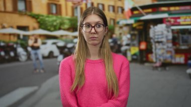 Pembe kazaklı kadın, caddede vitrin ve park halindeki arabaların yanında yan yan bakıyor..